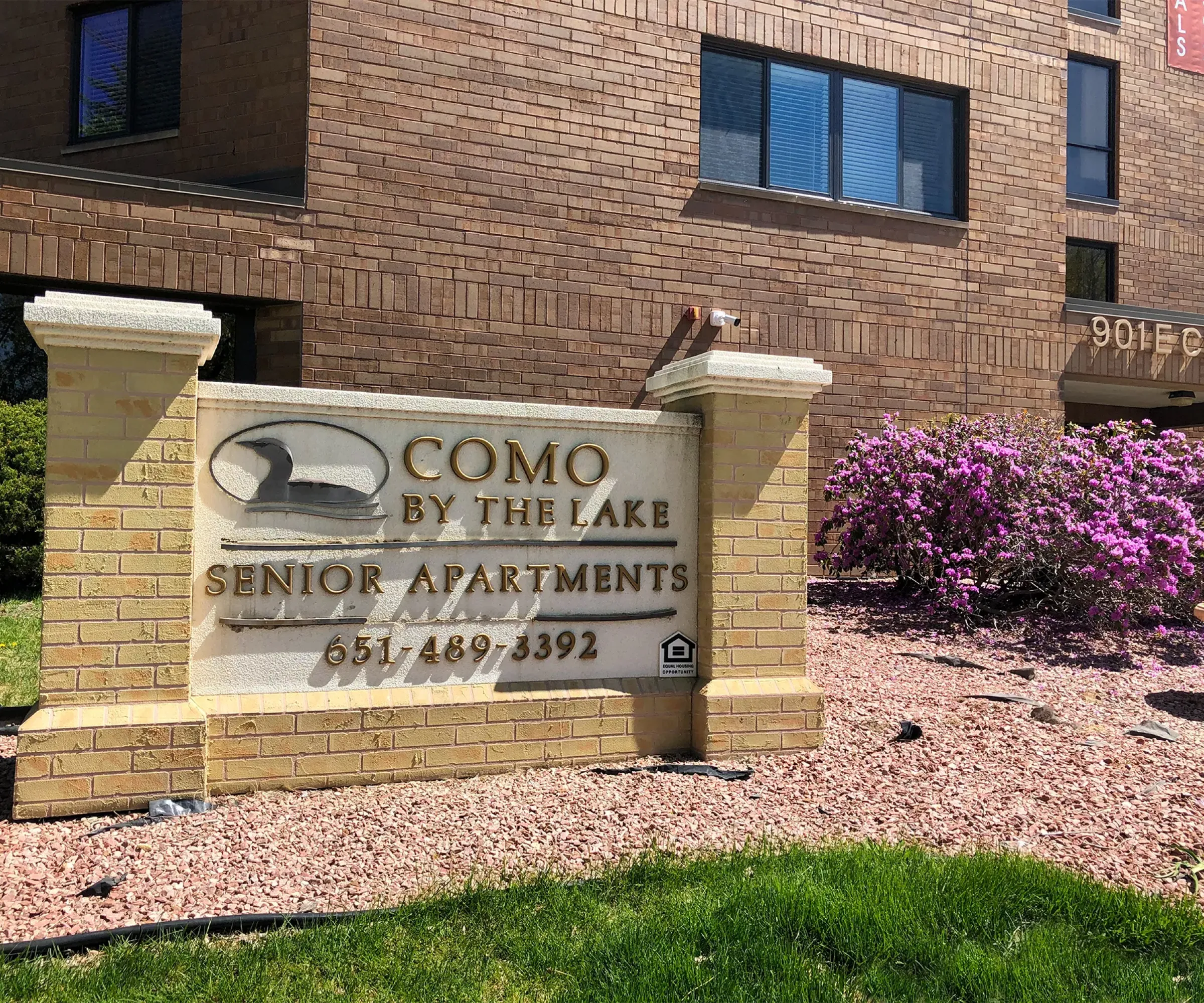 Aeon Affordable Homes and Community Sign for "Como By The Lake Senior Apartments," highlighting affordable apartment homes with flowers and a brick building in the background.