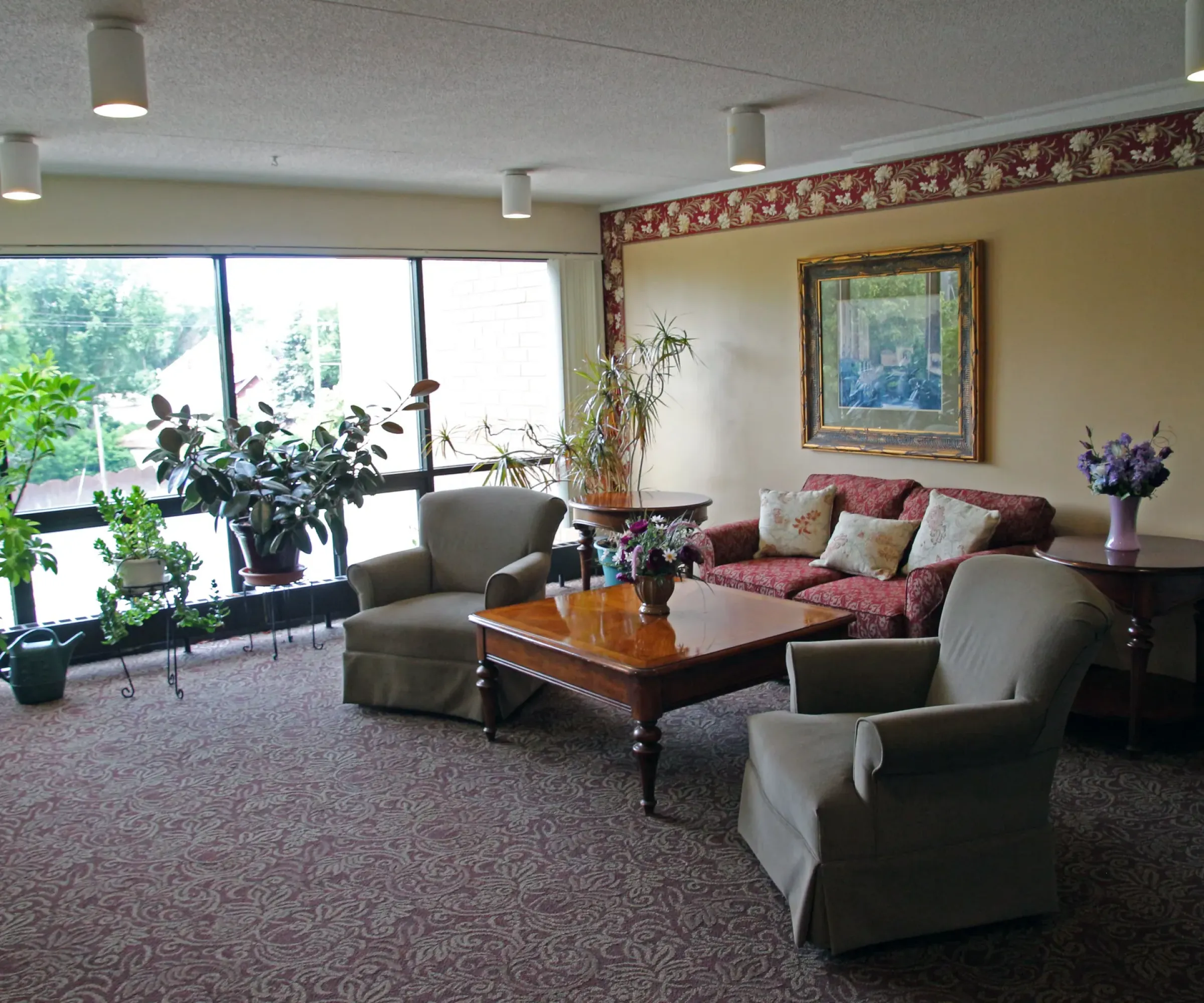 Aeon Affordable Homes and Community Cozy living room in affordable apartment homes with large window, potted plants, chairs, a sofa, and a wooden coffee table. Framed painting on wall.